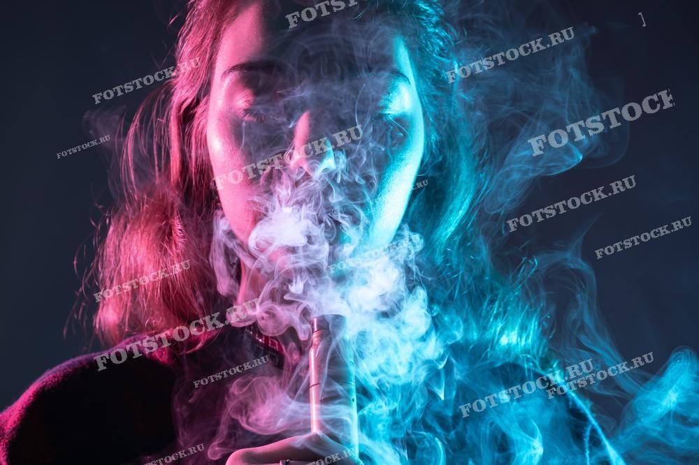 Фотография изображает молодую женщину, вдыхающую дым от электронной сигареты. Лицо частично скрыто клубами дыма, создавая мистический эффект. Эта фотография является иллюстрацией современной культуры вейпинга.